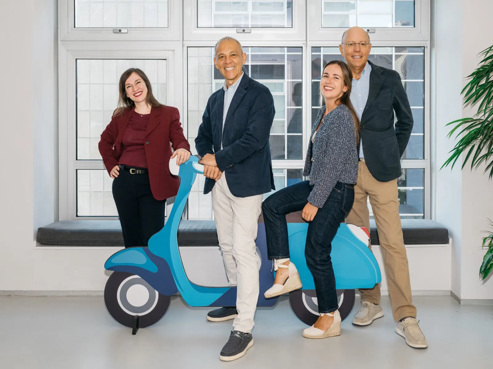 Las cuatro personas del equipo de HeliOx sonríen junto a una Vespa azul de cartón delante de un ventanal; una de ellas va sentada en el asiento